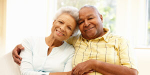 Two senior citizens smiling together as they hug while sitting on a couch.